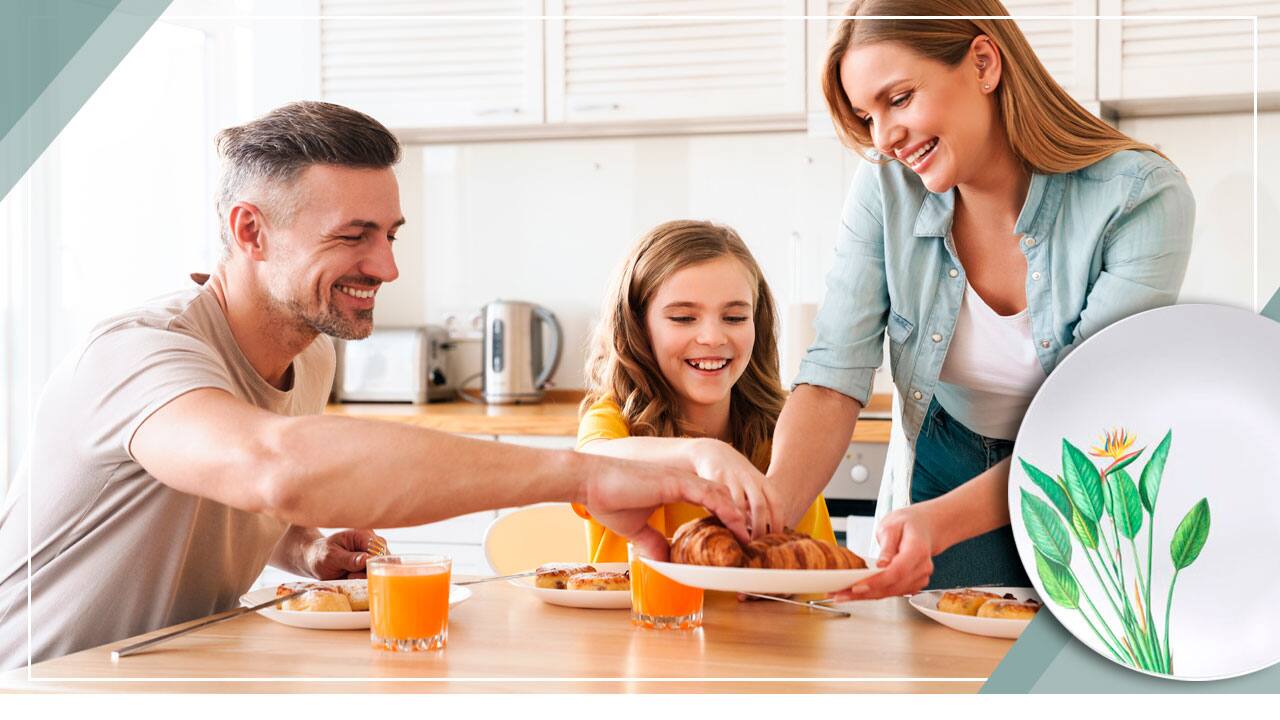 família casal homem rapaz jovem filha menina garota mulher moça servindo alimento salgado sorrisos sorrindo alegria alegres cozinha mesa marrom bege copos suco laranja pratos brancos branco estampa floral faca talher talheres fundo jarra cafeteira equipamentos pratas prata cinza janela janelas branca brancas fechada fechadas