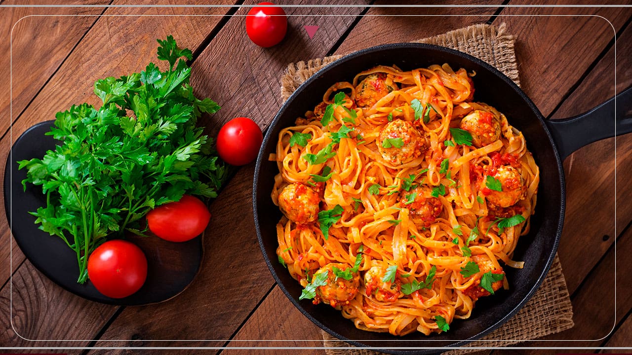 imagem frontal frente macarrão molho tomate vermelho bolinhos carne tempero verde salsinha frigideira tomates verdura hortaliça bancada marrom madeira
