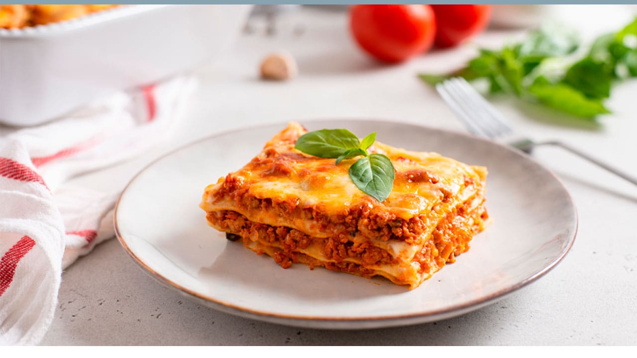 lasanha cortada pedaço carne massa folha verde prato pequeno branco mesa toalha garfo tomates vermelhos travessa refratário