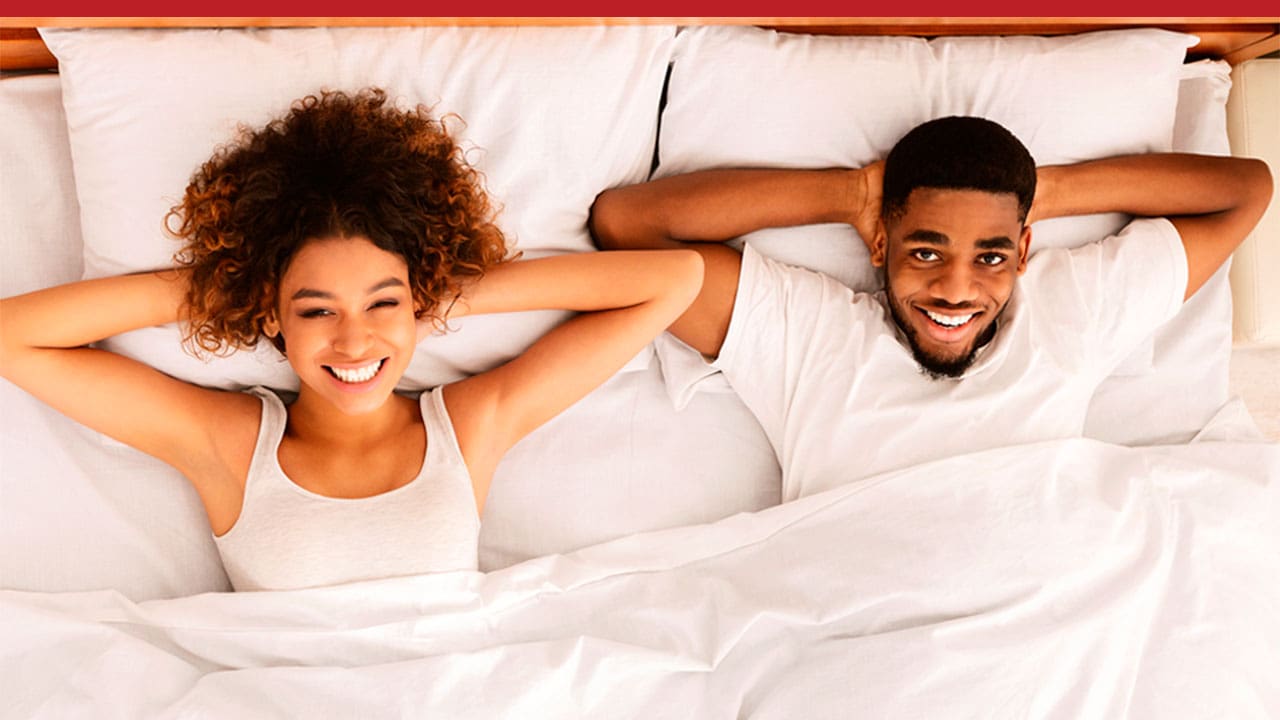 imagem mostra um casal sorridente deitados numa cama com roupas, travesseiros e lençóis brancos