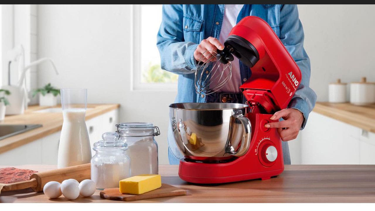 Batedeira Planetária Arno vermelha prata cinza brilho reflexo mãos pessoa em pé acionando botão vermelho camisa jeans azul camiseta branca potes transparentes pó branco farinha açúcar manteiga barra amarela ovos brancos rolo mesa bancada madeira marrom fundo torneira pia pequeno vaso branco planta folha verde janela pote