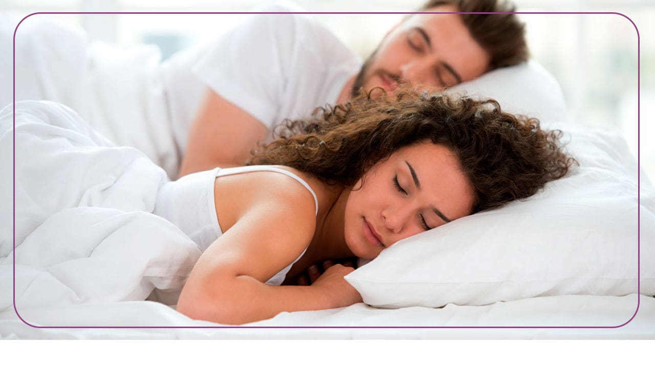 imagem mostra um casal dormindo em uma cama confortável com lençóis e travesseiros brancos