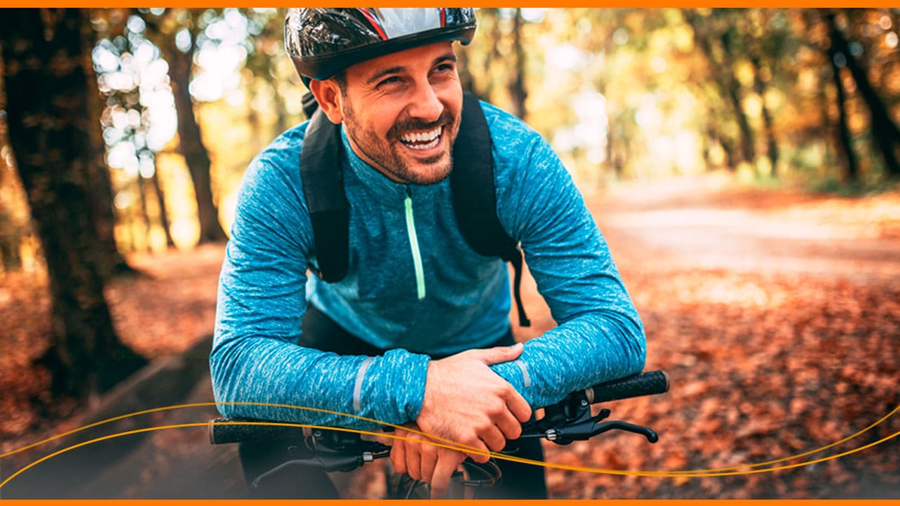 homem rapaz jovem sorrindo sorriso feliz barba capacete mochila preta blusa azul verde mãos bicicleta campo trilha estrada natureza árvores folhas chão