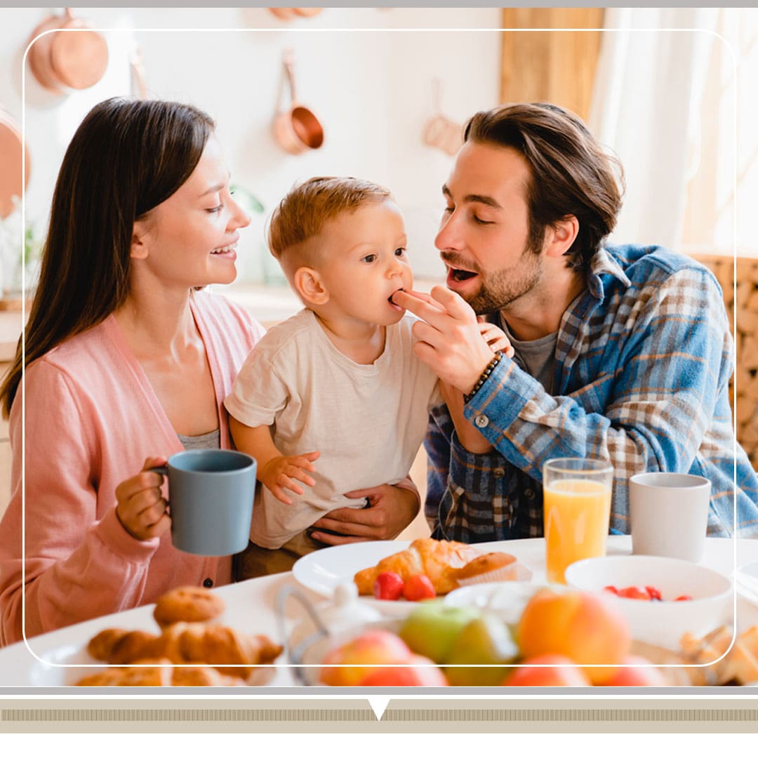 família reunida mãe pai filho sorrisos sorrindo mulher moça jovem mão xícara azul nenê bebê criança pequena menino homem moço rapaz dedo boca alimentando alimentação café mesa cheia fundo decoração panelas frigideiras parede