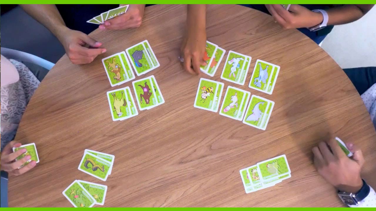 imagem mostra uma mesa com pessoas ao redor jogando o jogo do mico