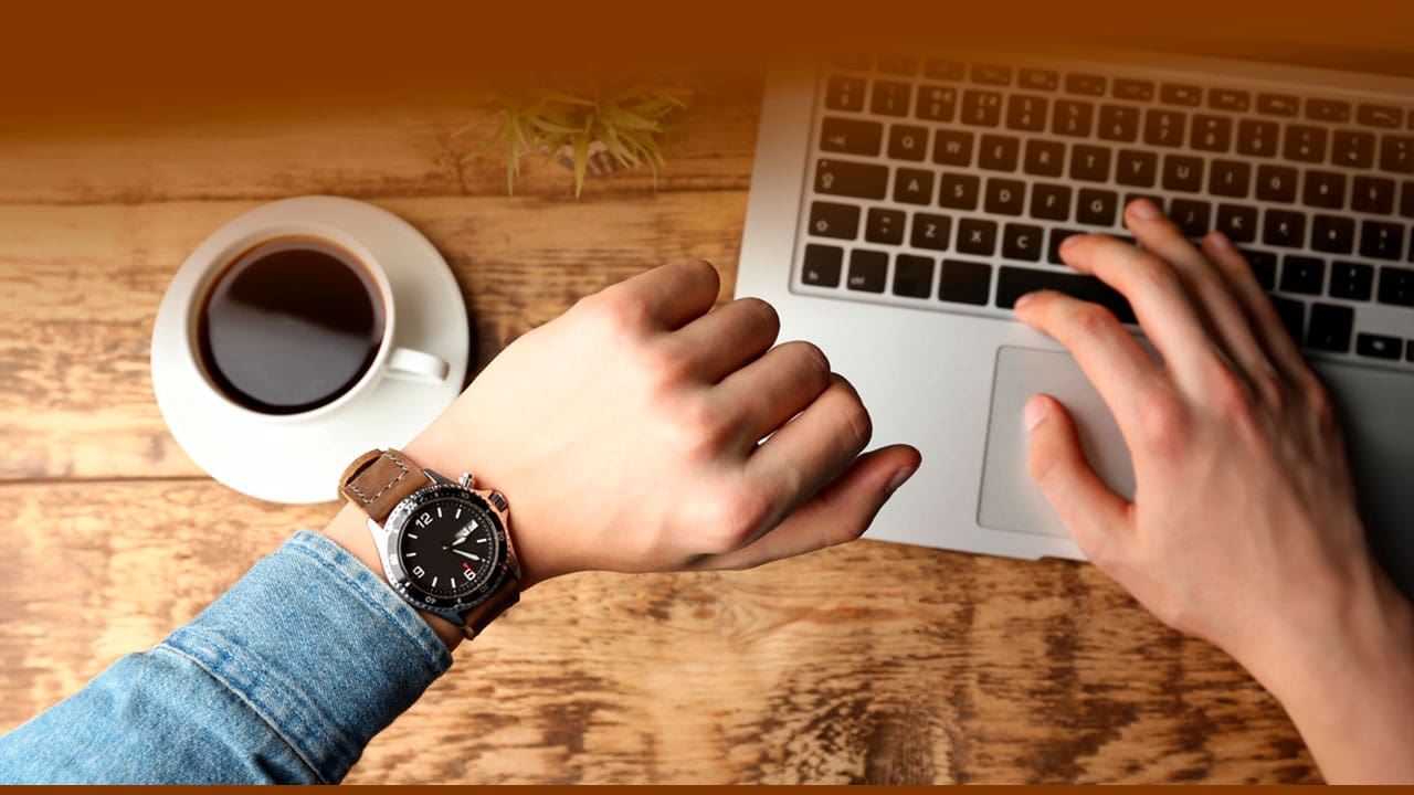 mãos masculinas homem relógio pulso marrom analógico camisa jeans azul notebook prata cinza teclas pretas teclado preto xícara café vaso pequeno planta folha verde bancada mesa madeira