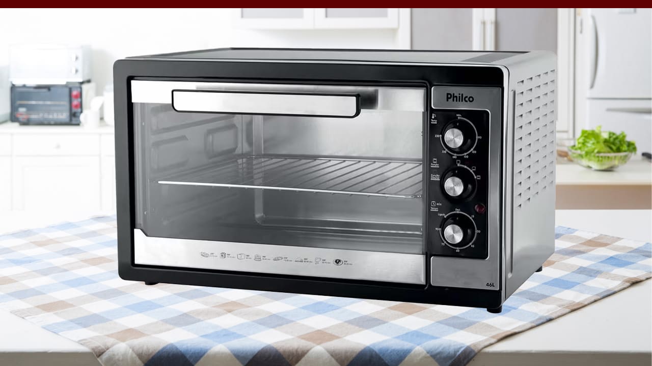 imagem frontal frente Forno Elétrico Philco Multifunção prata cinza preto três botões tampa porta fechada bancada branca toalha mesa quadriculada quadrados colorida fundo armário branco verdura verde pote