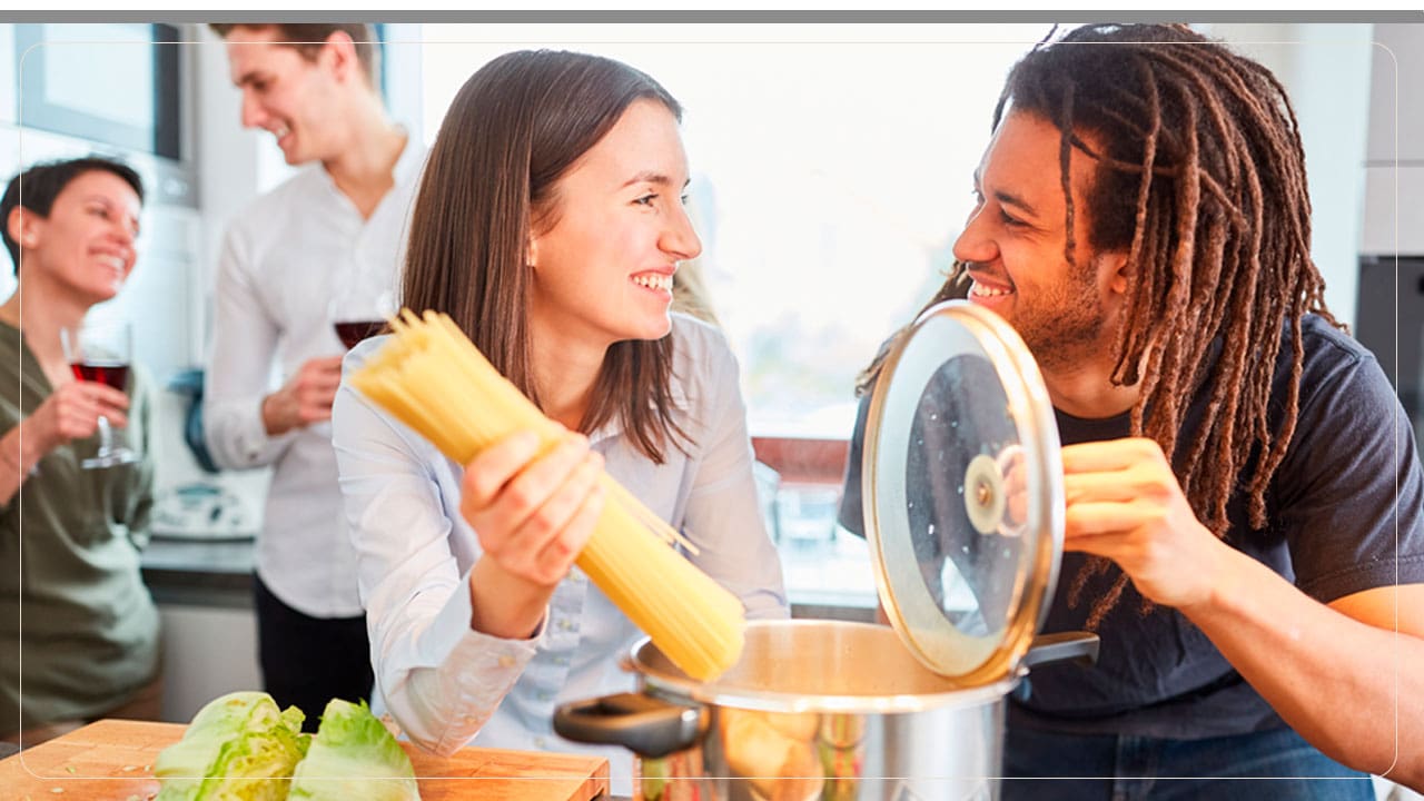 Um casal colocando macarrão na panela para cozinhar e outro casal ao fundo bebendo vinho