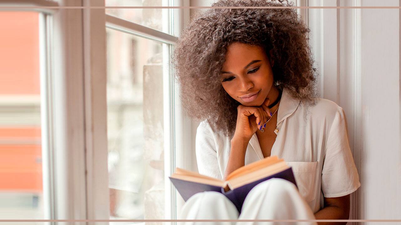 Mulher sentada próximo a janela lendo um livro
