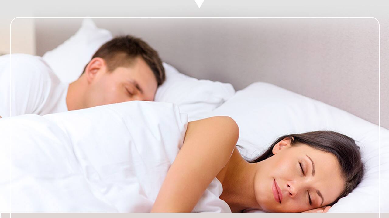 imagem mostra um casal dormindo em uma cama confortável com lençóis e travesseiros brancos