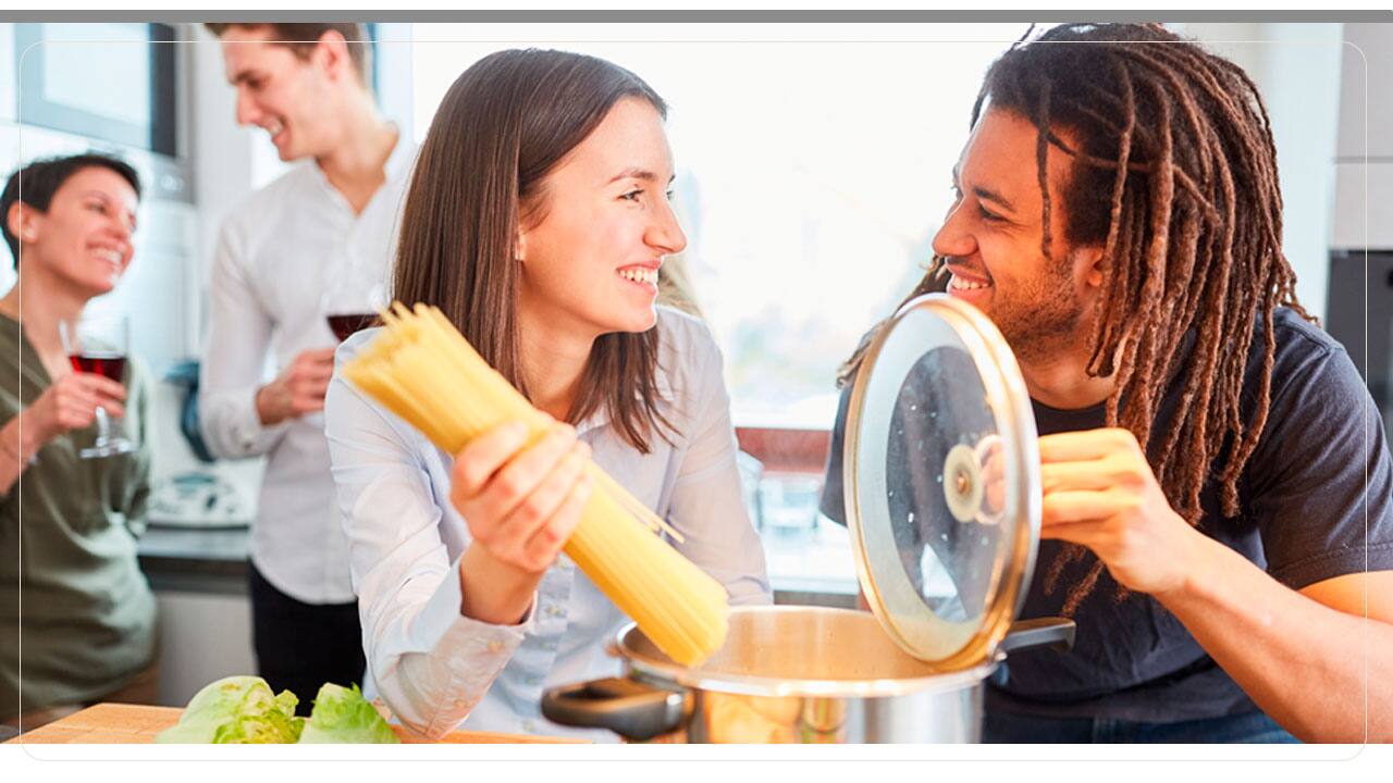 casais amigos jovens pessoas sorrindo sorrisos moços rapazes moças mulheres homens panela tampa macarrão mãos tábua marrom legumes vegetais cortados taças vinho bebida 